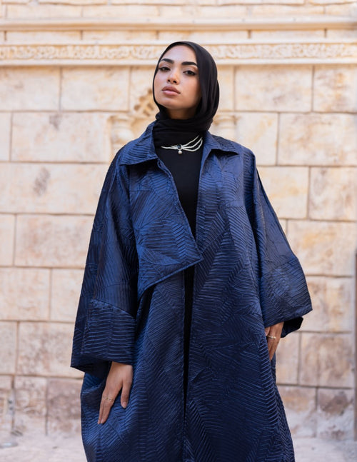 Woman wearing a blue textured coat and black hijab standing against a beige brick wall.