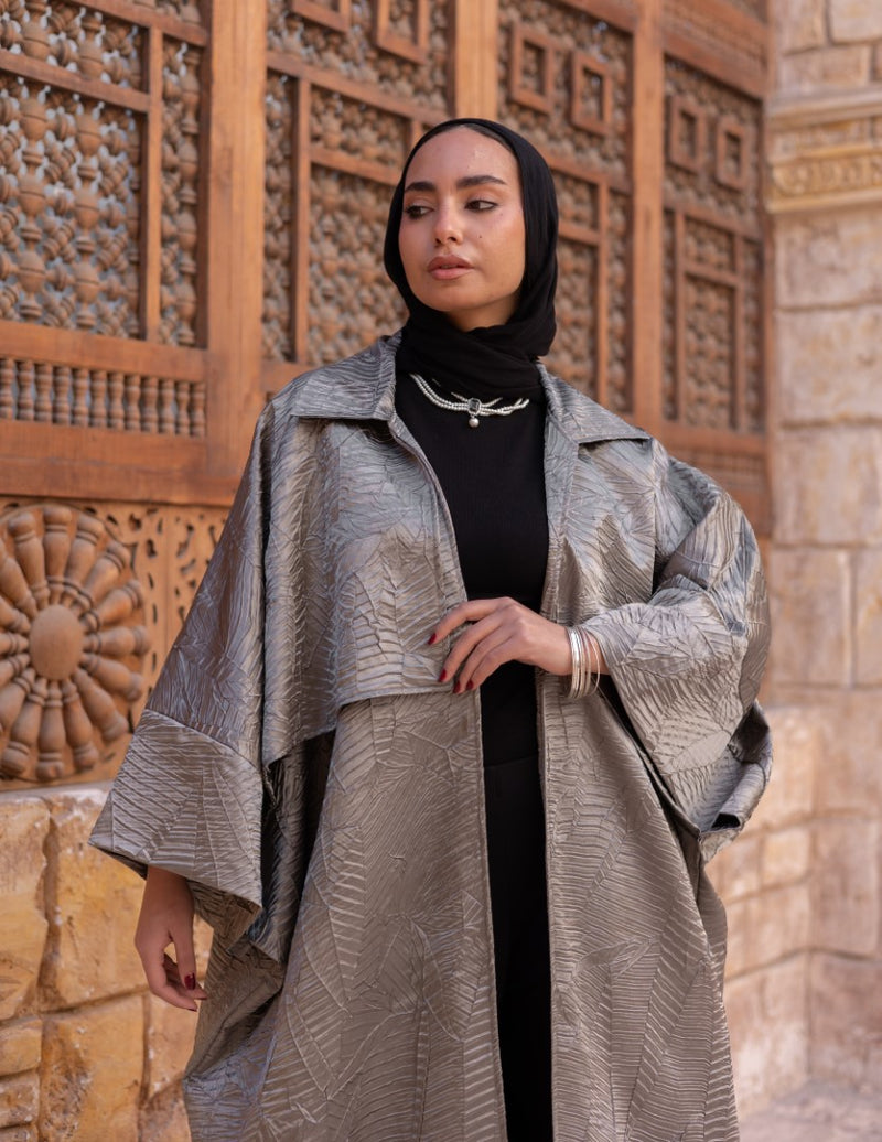 Woman wearing a textured silver coat and black hijab standing in front of a stone wall with wooden carvings.