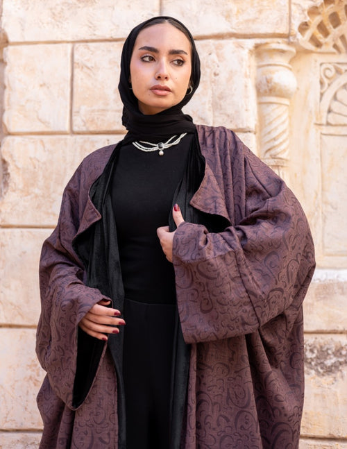 Woman wearing a dark purple patterned coat over a black outfit against a stone wall.