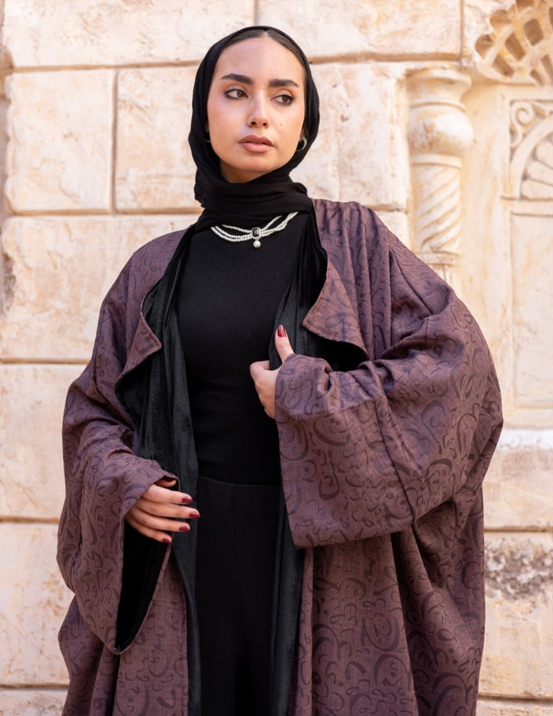 Woman wearing a dark purple patterned coat over a black outfit against a stone wall.