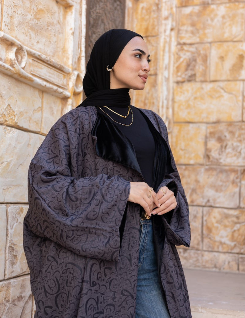 Woman wearing a dark patterned coat and black hijab against a stone wall.