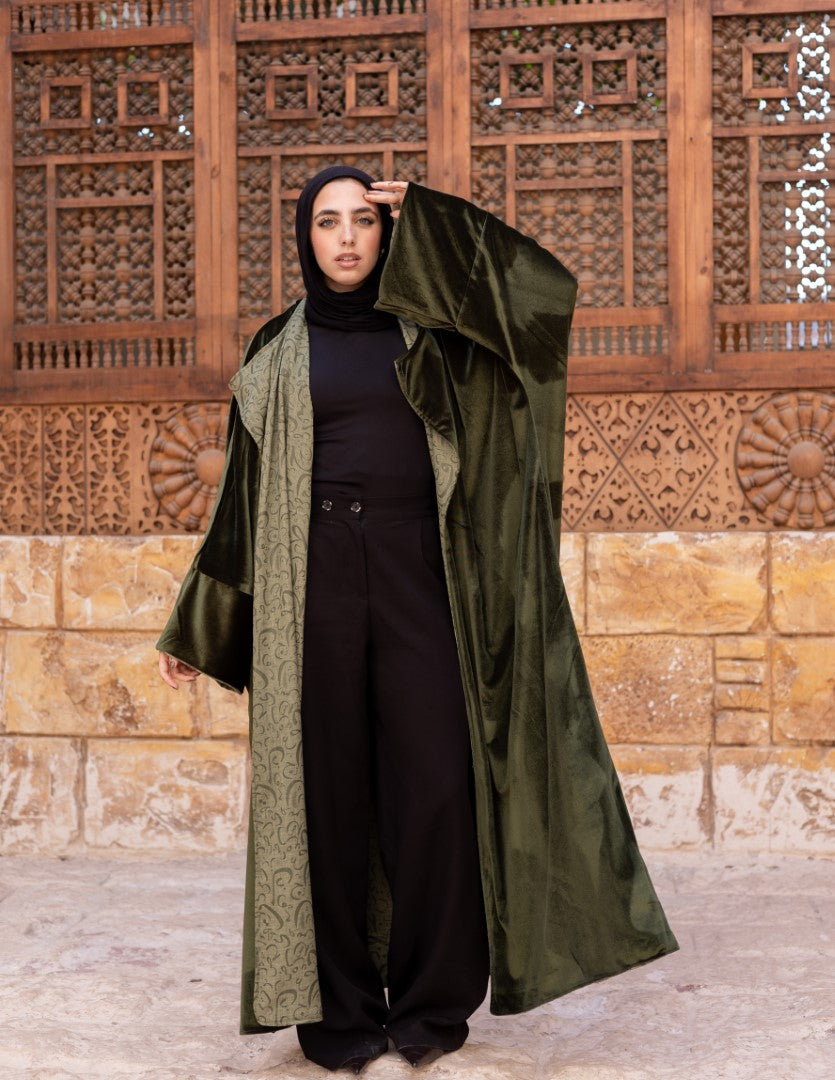 Person wearing a green coat over a black outfit in front of a decorative stone wall.