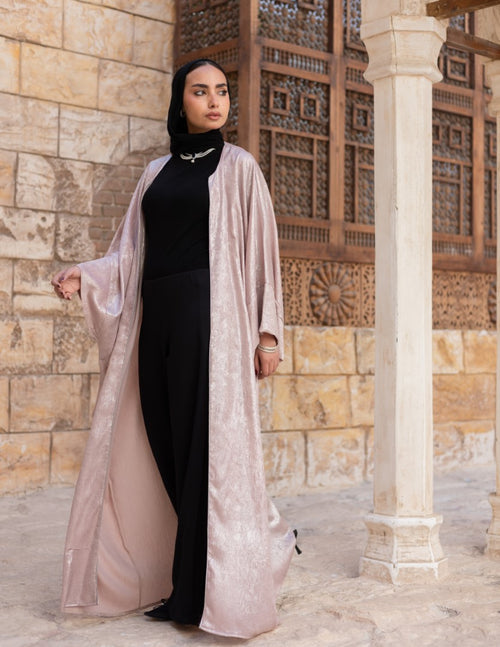 Woman wearing a black outfit with a beige abaya in an outdoor setting with stone walls.