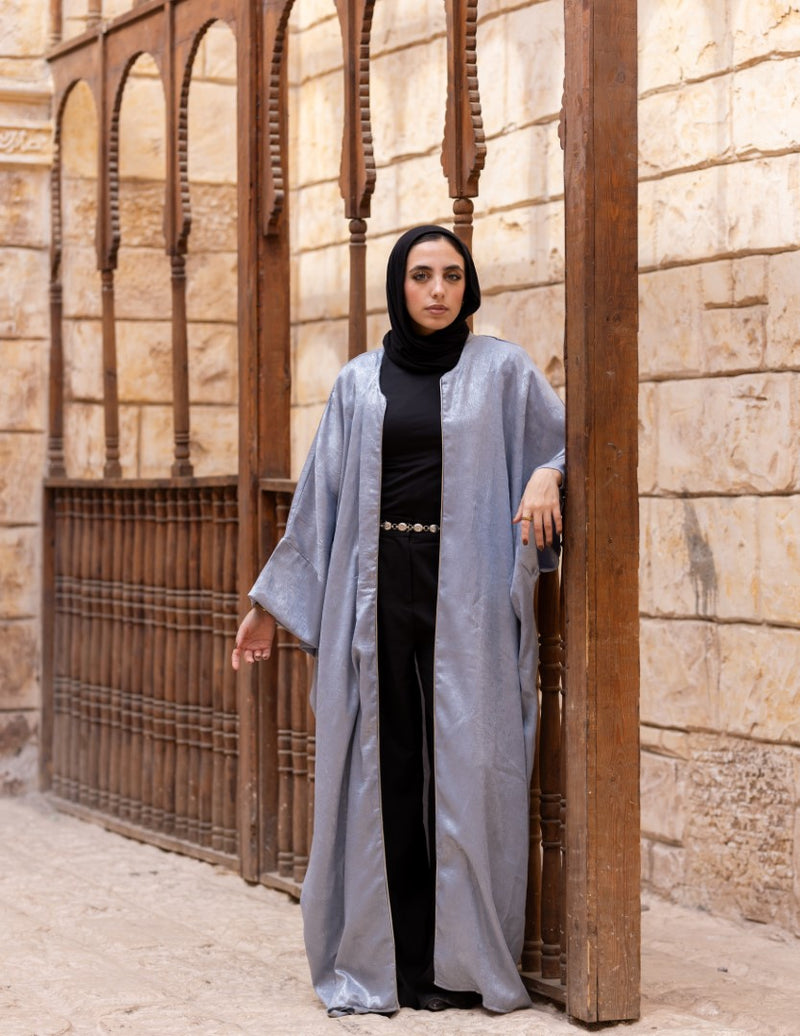 Woman in a long light gray coat and black hijab standing in front of stone walls.