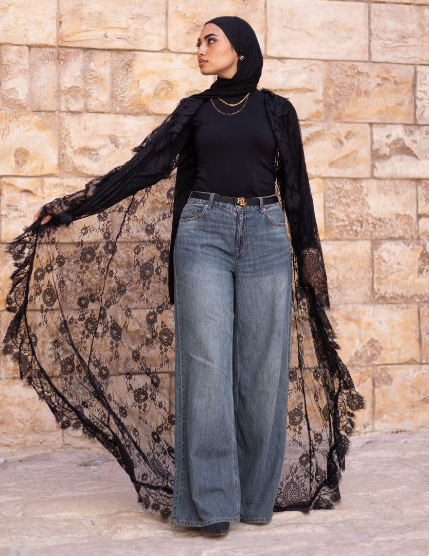 Woman wearing a black lace dress over a black top and blue jeans against a stone wall.