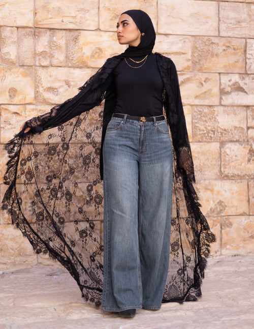 Woman wearing a black lace dress over a black top and blue jeans against a stone wall.