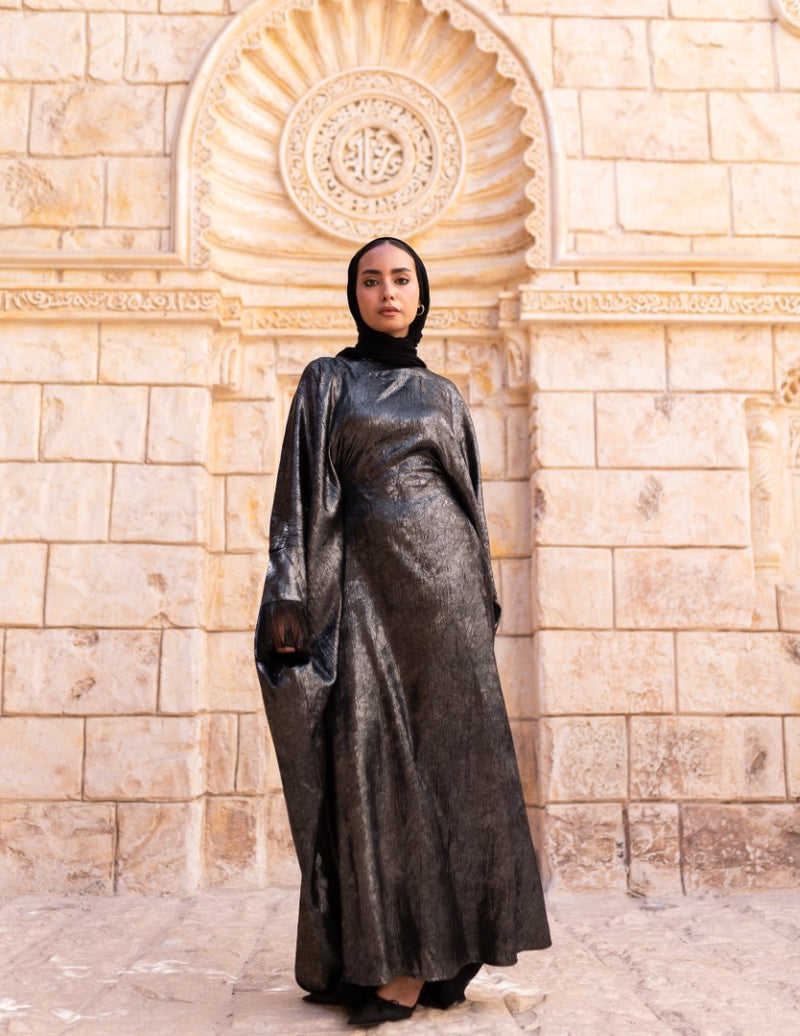 Woman in a long, dark-colored dress standing in front of a stone wall with an archway.